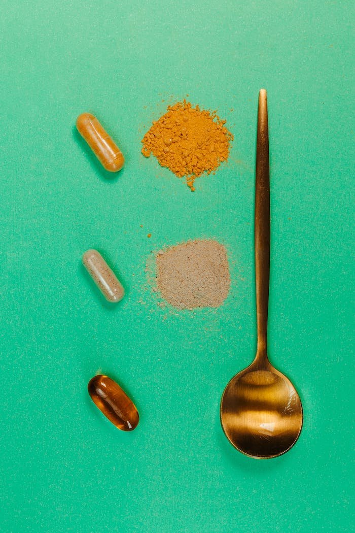 Top view of a golden spoon with capsules and powders on a vibrant green surface, representing supplements.